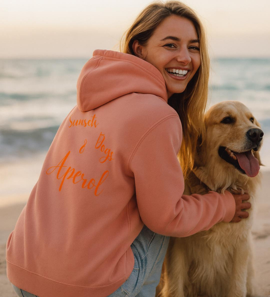 Frau im rosafarbenen Hoodie mit Aufschrift „Sunset • Dogs • Aperol“ sitzt lachend mit ihrem Golden Retriever am Strand bei Sonnenuntergang.