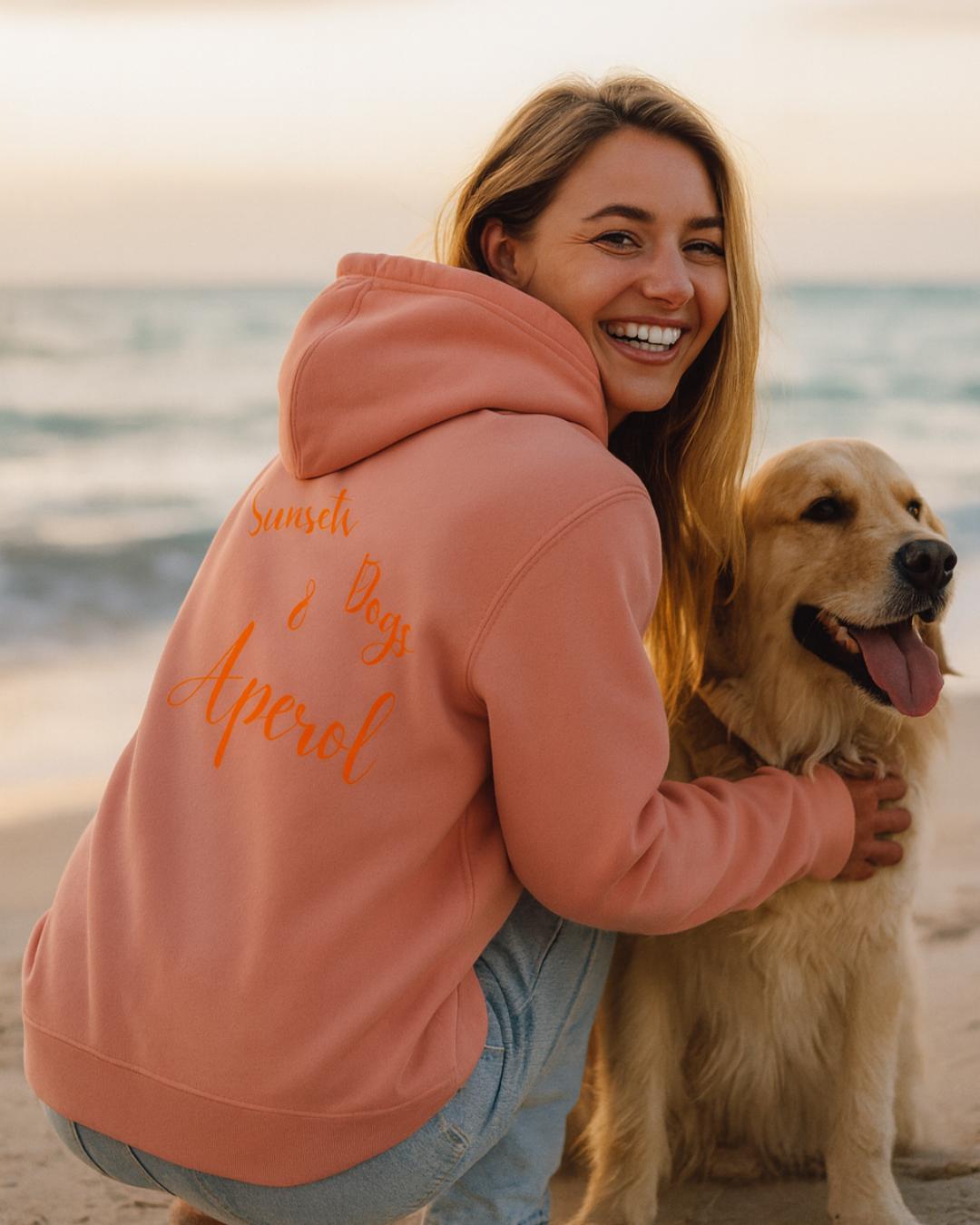 Frau im rosafarbenen Hoodie mit Aufschrift „Sunset • Dogs • Aperol“ sitzt lachend mit ihrem Golden Retriever am Strand bei Sonnenuntergang.