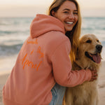 Frau im rosafarbenen Hoodie mit Aufschrift „Sunset • Dogs • Aperol“ sitzt lachend mit ihrem Golden Retriever am Strand bei Sonnenuntergang.